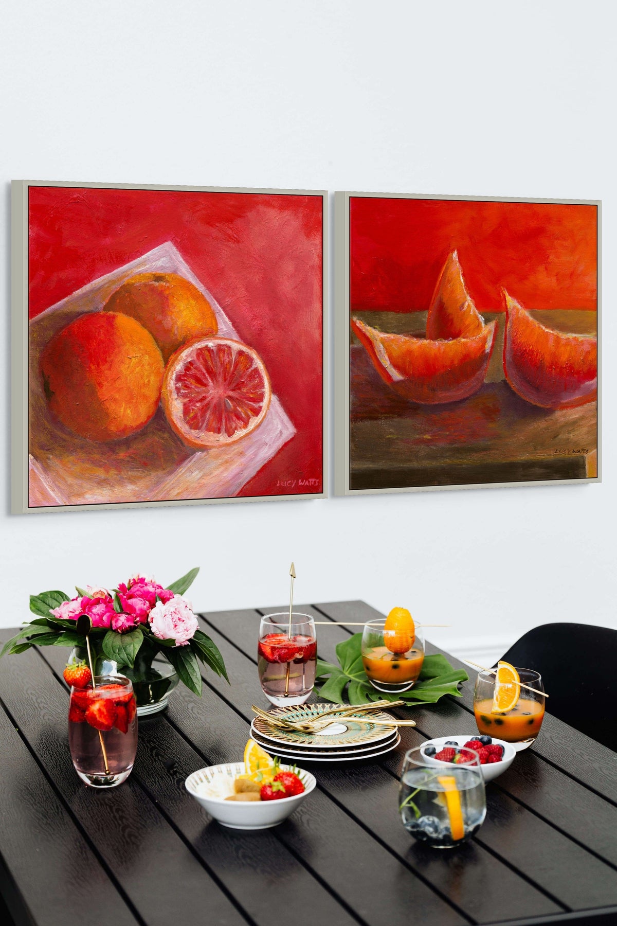 Two framed prints of fruity paintings by Australian artist Lucy Watts of bright fruit on a table with a floral arrangement and drinks. Colourful table setting.
