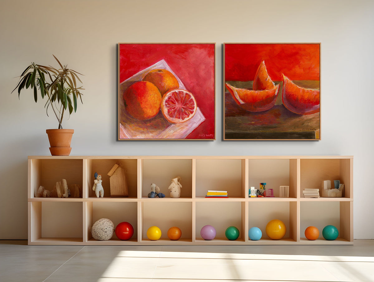Two abstract paintings of fruit on a wall above a wooden shelf with decorative items.